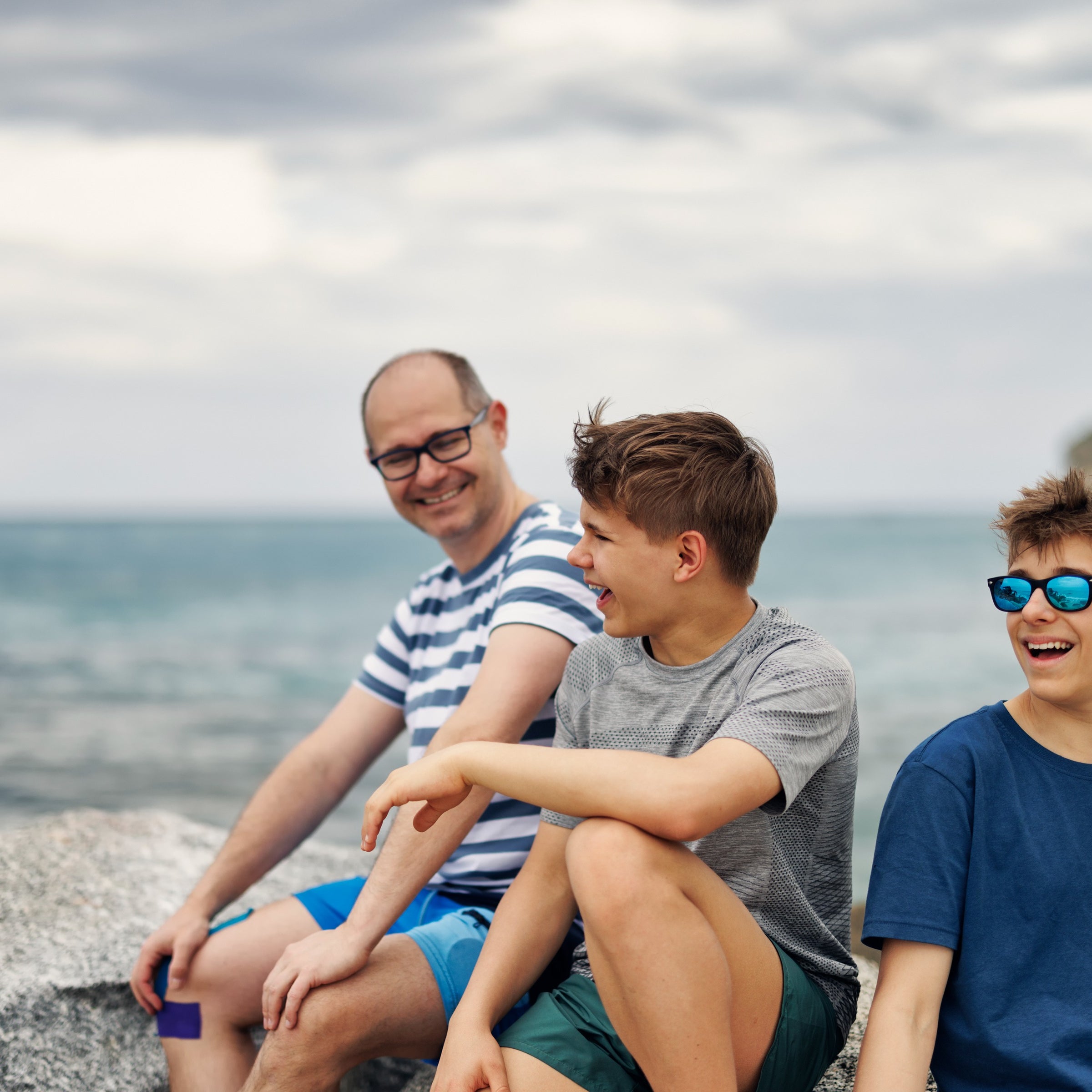 Three people are smiling and sitting on rocks by the sea.