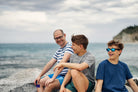 Three people are smiling and sitting on rocks by the sea.