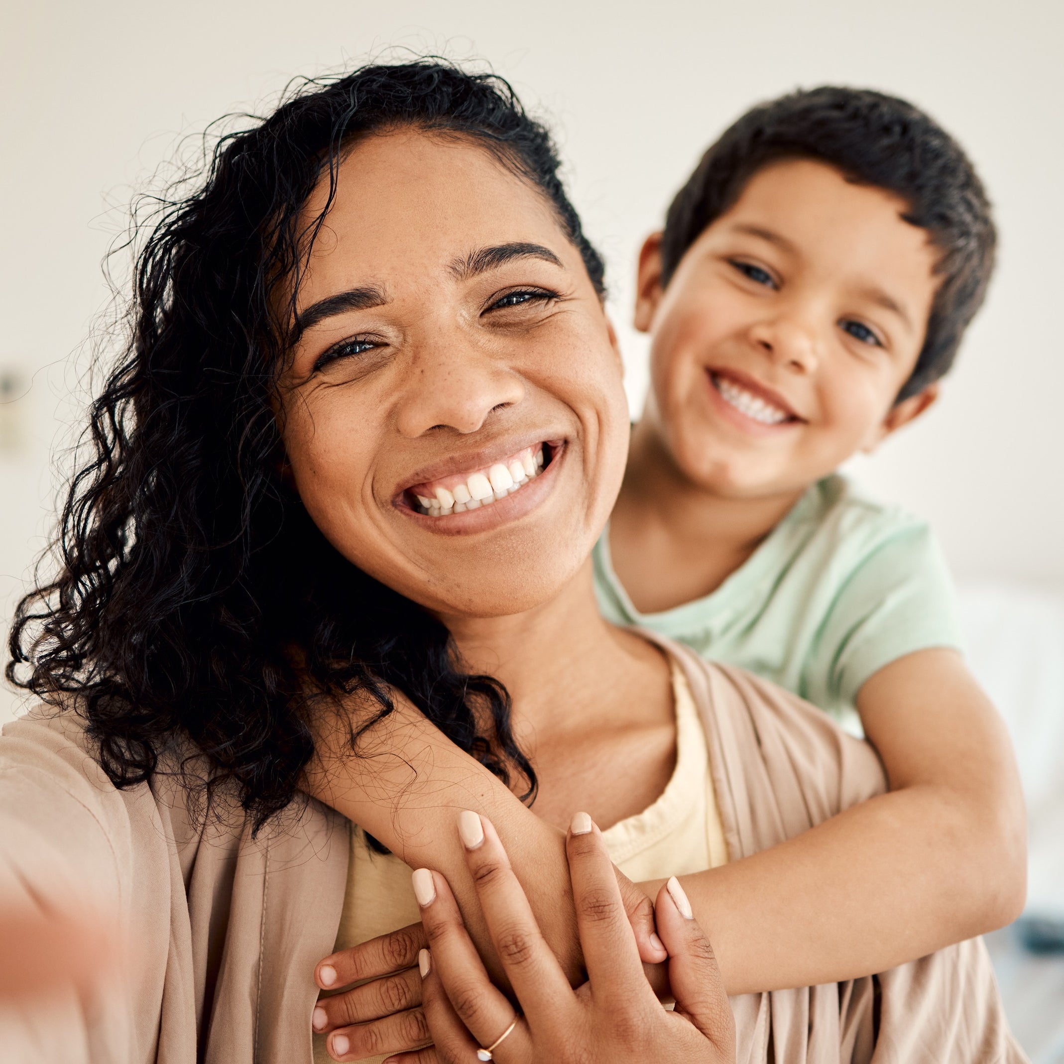 A smiling woman with curly hair holds a child, who hugs her from behind, both looking happy.