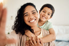 A smiling woman with curly hair holds a child, who hugs her from behind, both looking happy.