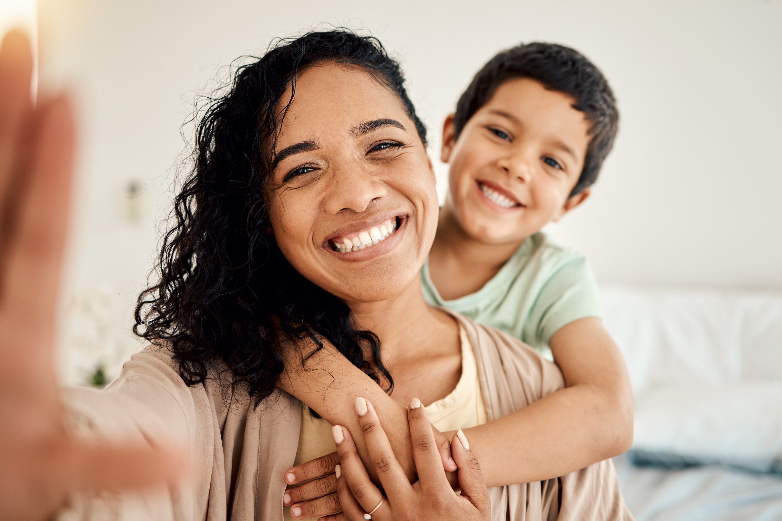 A smiling woman with curly hair holds a child, who hugs her from behind, both looking happy.